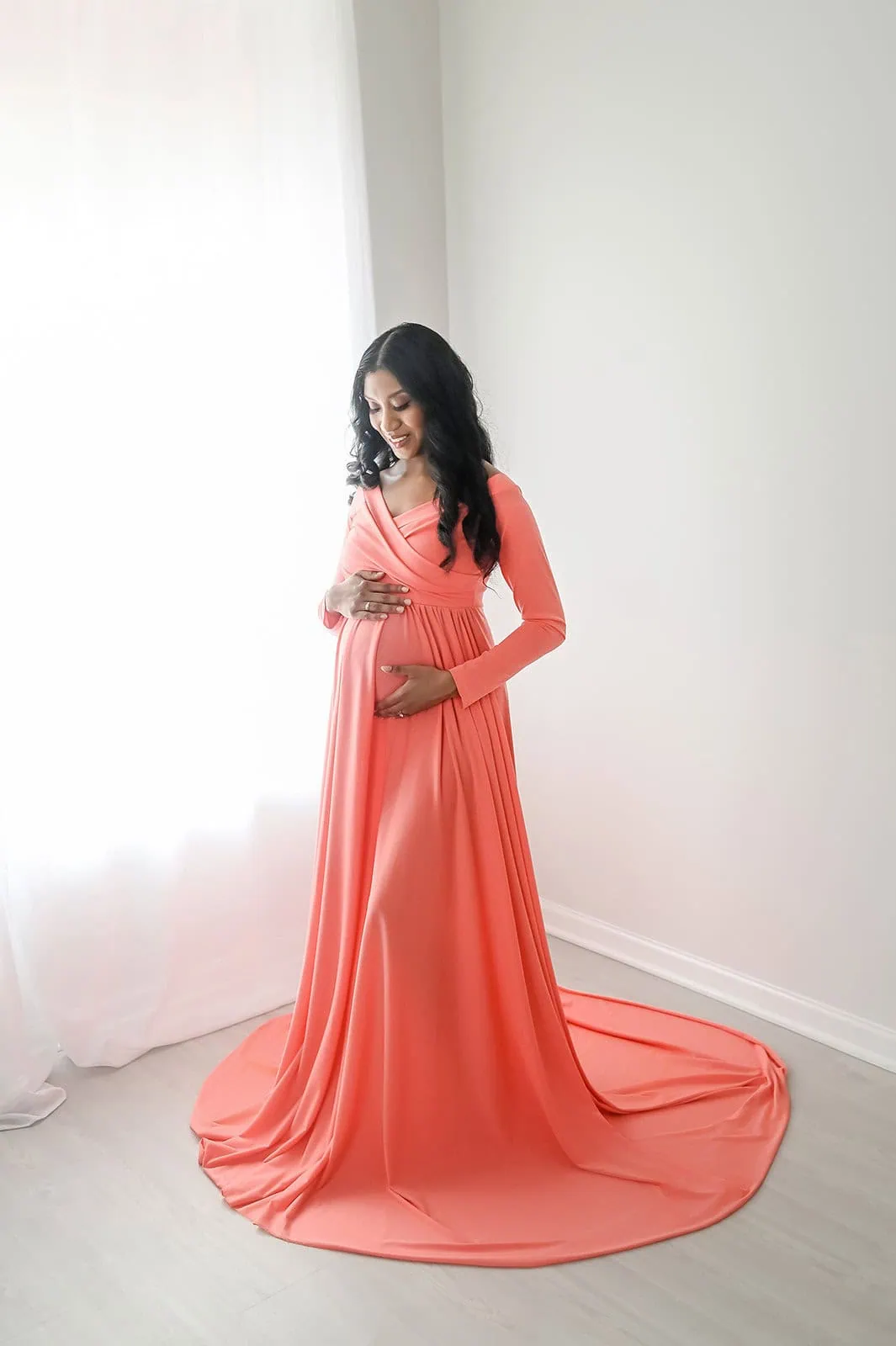 A pregnant woman stands indoors in a well-lit room, gently cradling her baby bump. She is wearing a long, flowing coral dress with long sleeves. The soft lighting and light-colored walls add a calm and serene atmosphere to the image. - Image by Kristin Milito Photography, Chicago IL.