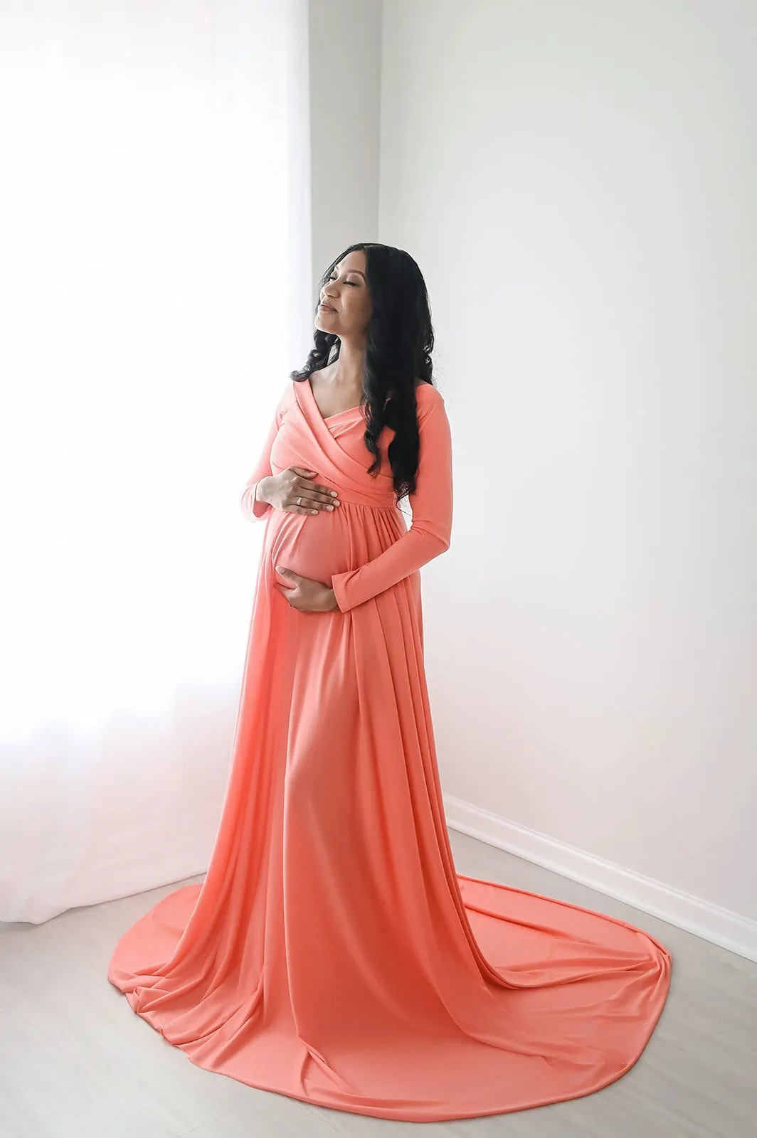 A pregnant woman stands in a softly lit room, wearing a long, flowing coral dress. She cradles her belly with both hands and has a serene expression on her face. The dress gently cascades to the floor, creating a graceful and elegant silhouette. - Image by Kristin Milito Photography, Chicago IL.