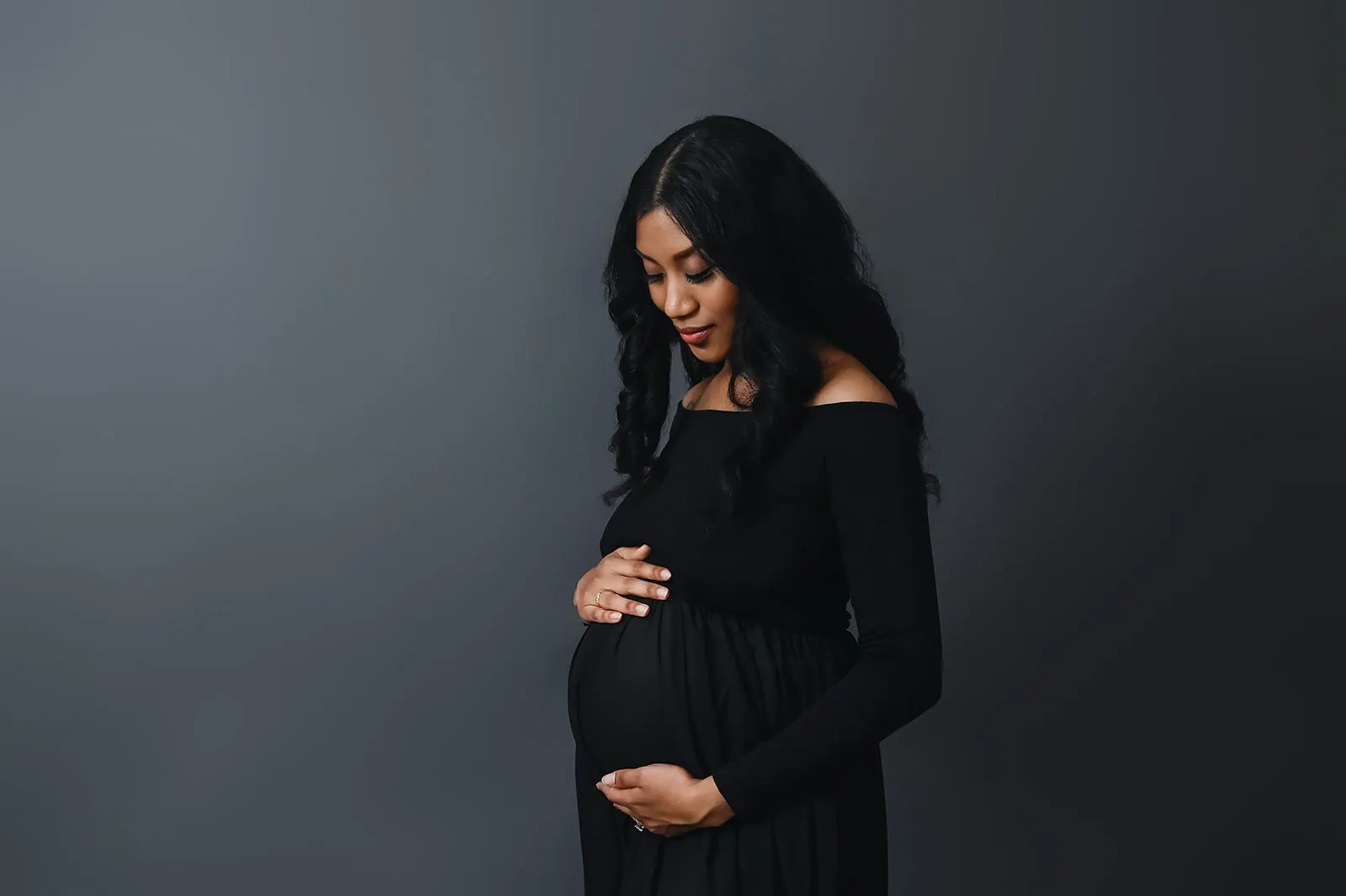 A pregnant woman in a black dress stands against a plain dark gray background. She is looking down and gently cradling her baby bump with both hands, exuding a calm and serene demeanor. - Image by Kristin Milito Photography, Chicago IL.