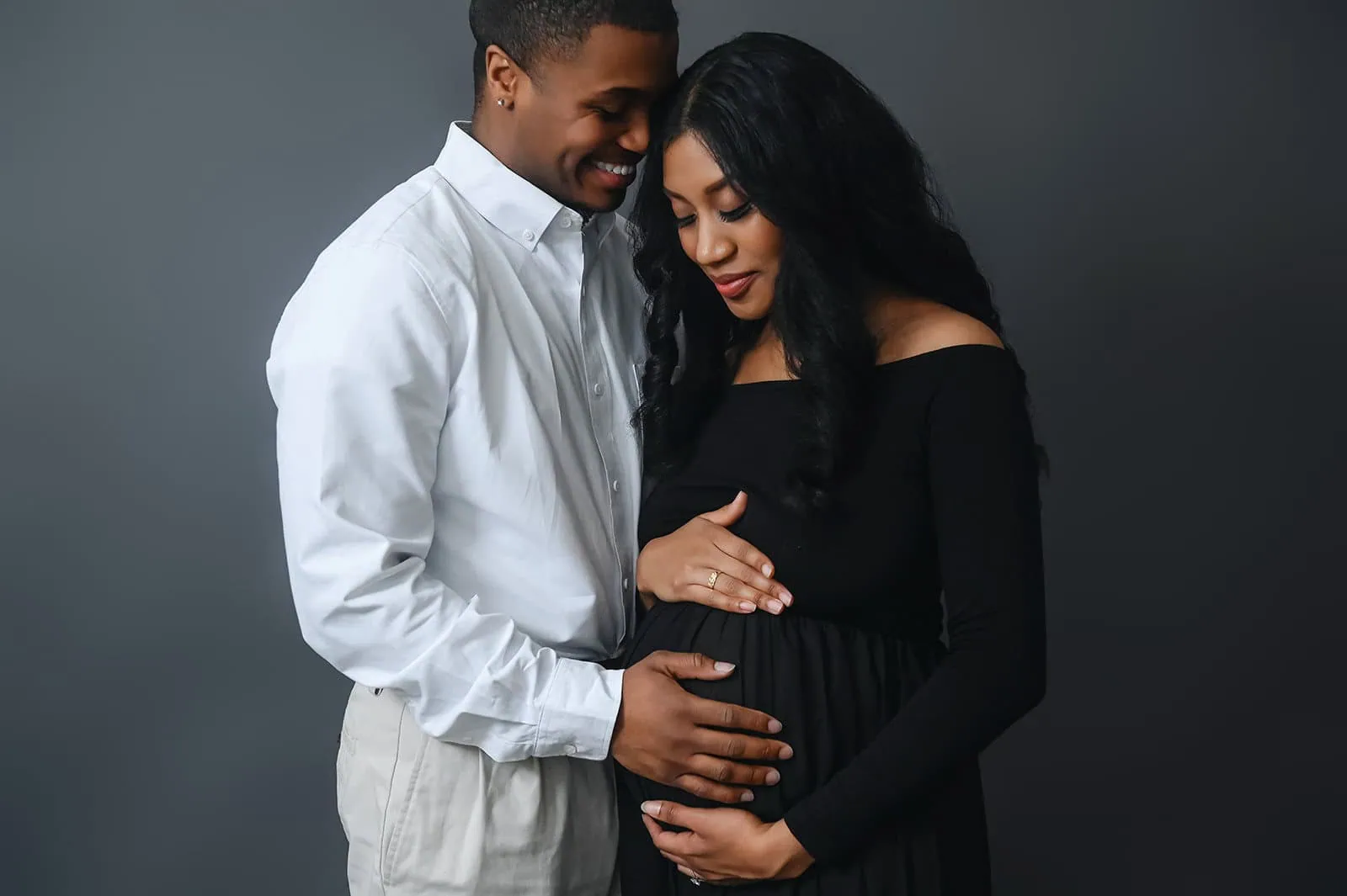 A couple stands closely together, with the woman in a black dress holding her baby bump. The man, wearing a white shirt and beige pants, gently places his hand on her belly and smiles warmly at her. The background is plain and dark. - Image by Kristin Milito Photography, Chicago IL.