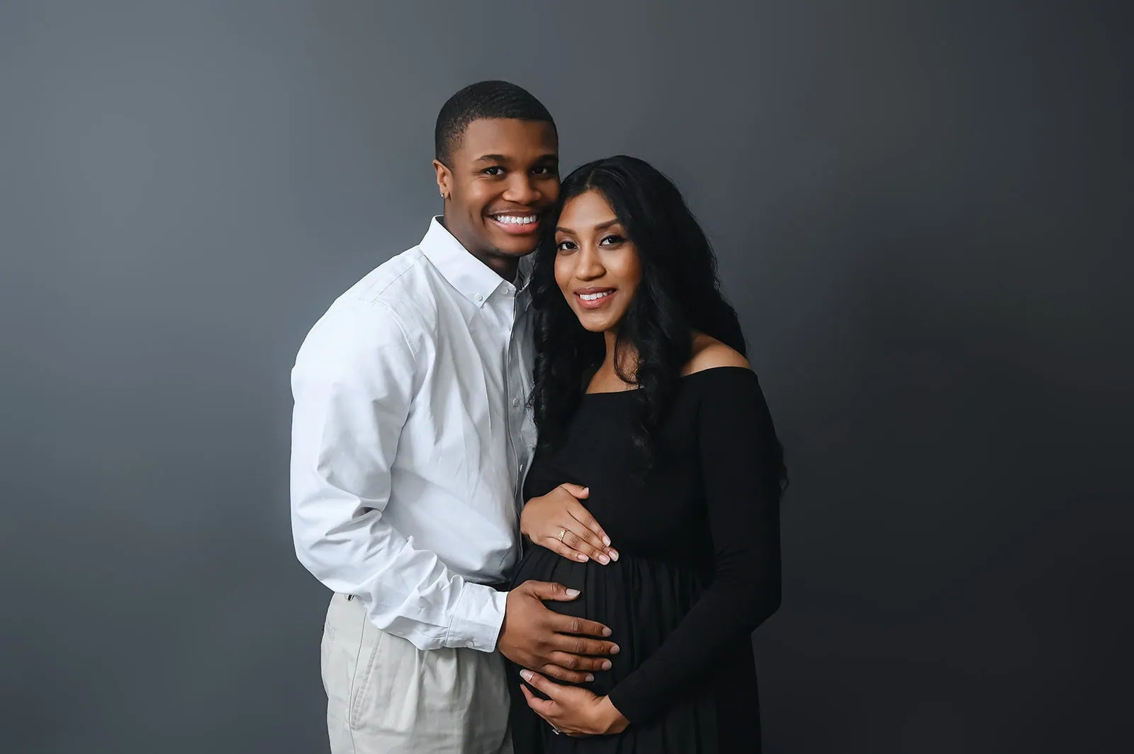 A smiling couple stands against a dark grey background. The man on the left, wearing a white shirt and light-colored pants, lovingly embraces the woman on the right, who is wearing a black off-the-shoulder dress and cradling her baby bump. - Image by Kristin Milito Photography, Chicago IL.
