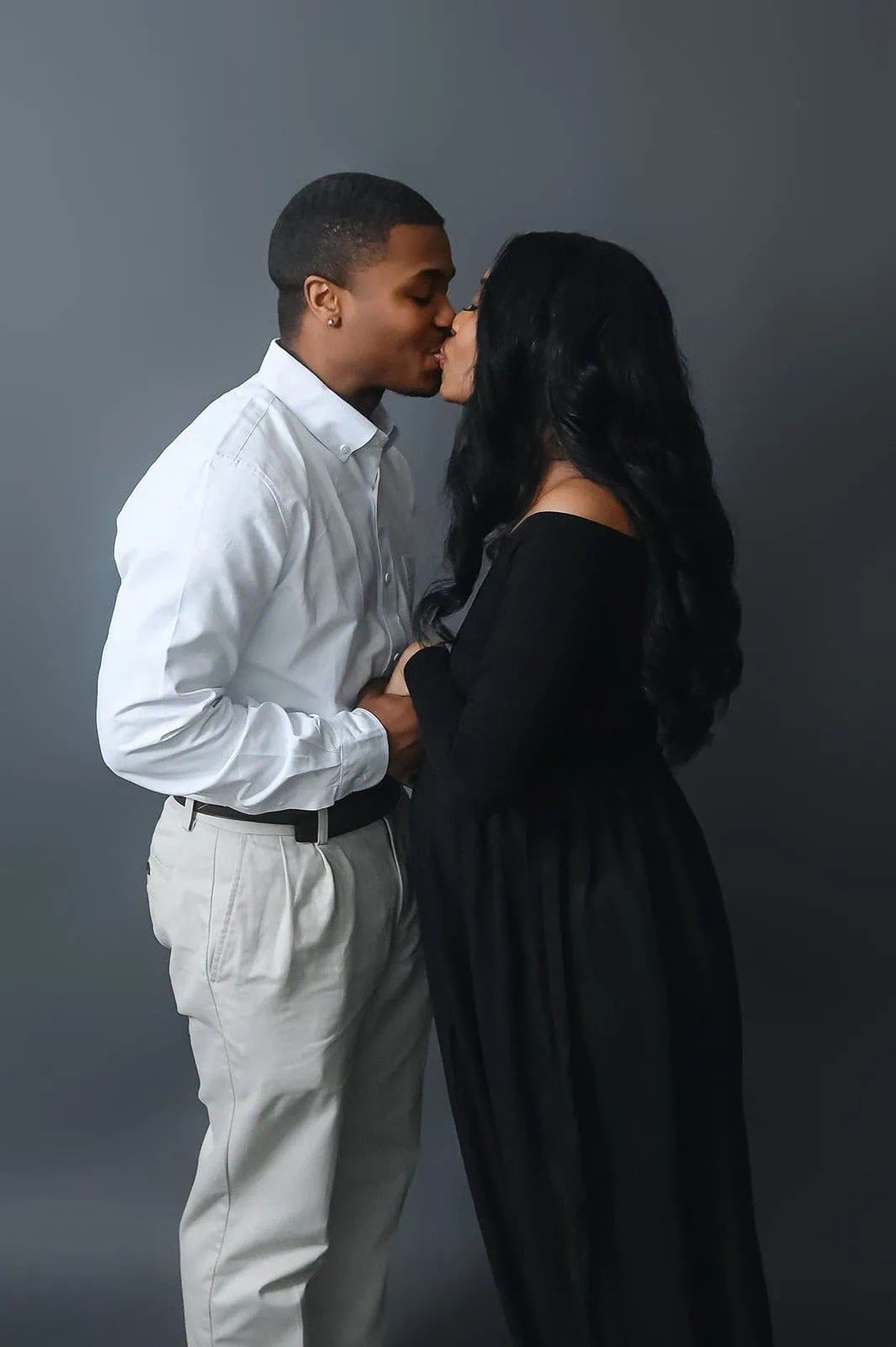 A couple stands against a gray backdrop, kissing tenderly. The man is wearing a white dress shirt and light-colored pants, while the woman is dressed in a long black dress. They hold hands, with the woman's hair cascading over her shoulders. - Image by Kristin Milito Photography, Chicago IL.