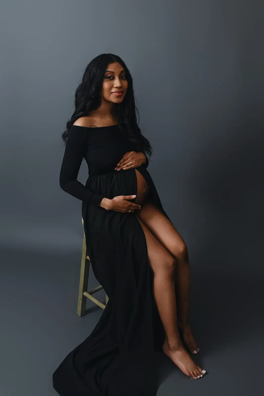 A pregnant woman wearing a long, flowing black dress is seated on a stool against a dark grey background. She has long hair and is cradling her bare belly with one hand while resting the other on her thigh. She is looking at the camera with a calm expression. - Image by Kristin Milito Photography, Chicago IL.