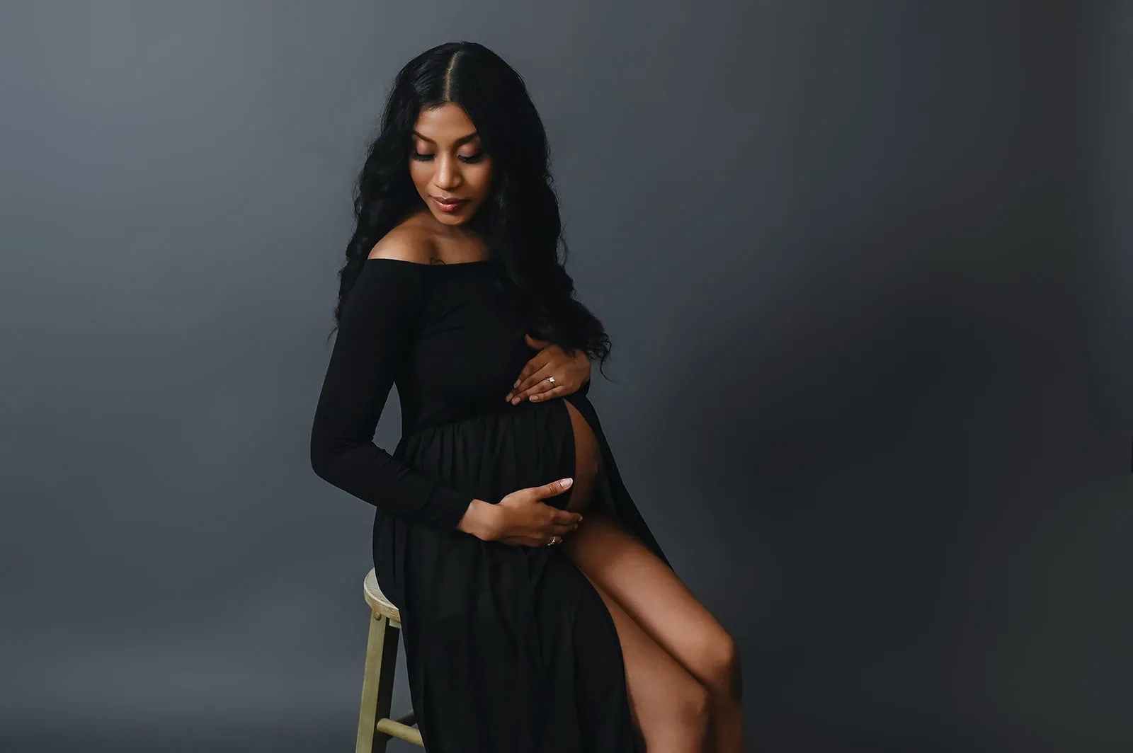 A pregnant woman with long, wavy hair sits on a stool against a dark gray backdrop. She is wearing a black, off-the-shoulder dress and is gently cradling her bare baby bump with her hands, looking down with a serene expression. - Image by Kristin Milito Photography, Chicago IL.