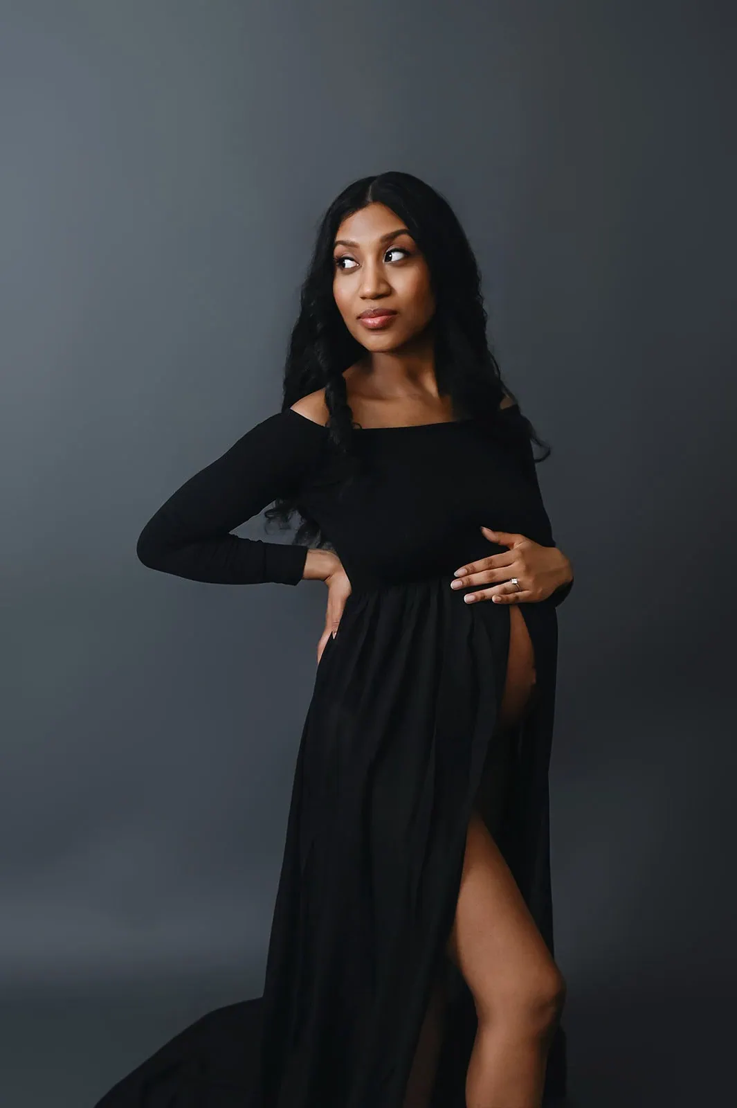 A pregnant woman with long black hair wearing a flowing black off-the-shoulder dress stands against a dark grey background. One hand is on her hip, and the other cradles her belly. She looks thoughtfully to the side. - Image by Kristin Milito Photography, Chicago IL.