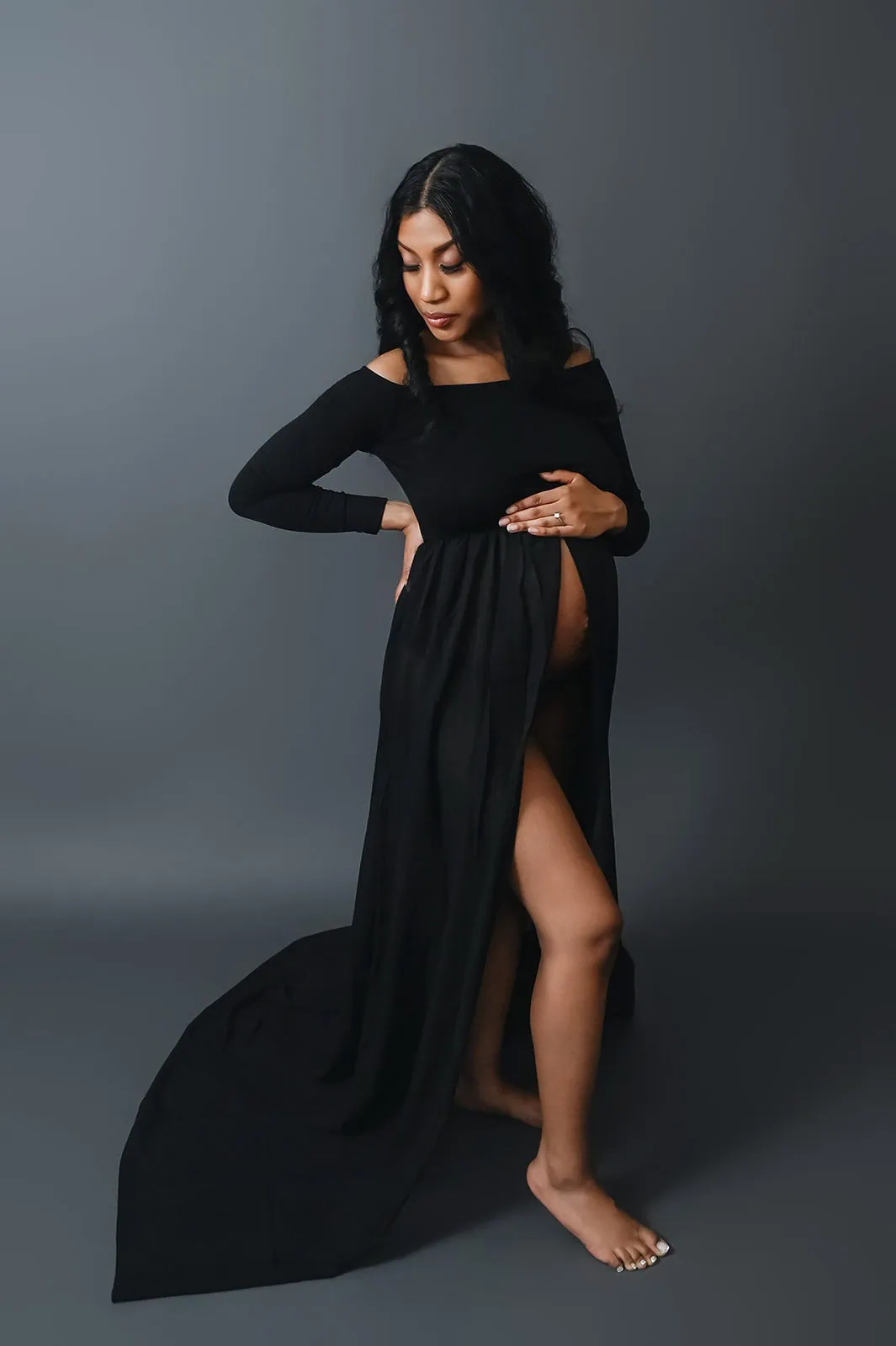 A pregnant woman stands barefoot against a dark gray background, cradling her bare belly with her left hand. She is wearing a long, black, off-the-shoulder gown with a high slit that reveals her right leg. Her gaze is cast downward. - Image by Kristin Milito Photography, Chicago IL.