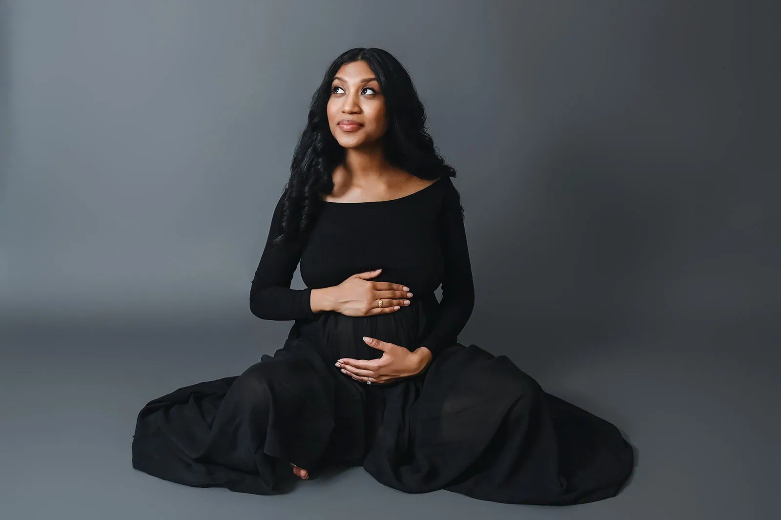 A pregnant woman with long, dark hair sits on the floor against a plain gray background. She wears a black, long-sleeved top and a flowing black skirt, cradling her belly with both hands and looking upwards with a serene expression. - Image by Kristin Milito Photography, Chicago IL.