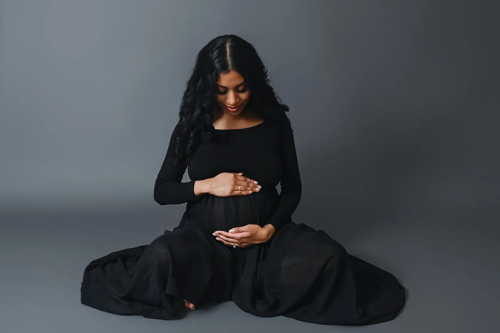 A pregnant woman with long, dark curly hair sits cross-legged on the floor against a plain gray background. She is wearing a black long-sleeved dress and gently cradles her baby bump with both hands while looking down, smiling softly. - Image by Kristin Milito Photography, Chicago IL.
