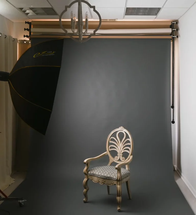 A photography studio setup featuring a single ornate chair with a patterned cushion placed on a gray backdrop, creating a cozy, home-inspired ambiance. A large softbox light is positioned on the left side, illuminating the scene. A ceiling light fixture is visible at the top of the image.