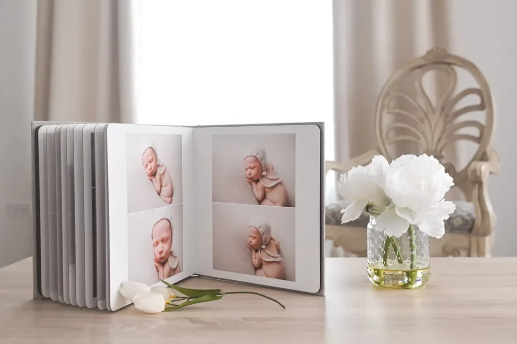An open photo album on a wooden table displays four pictures of a sleeping newborn. Next to the album is a vase with white flowers, and a single white tulip lies on the table. A decorative wooden chair adds a cozy touch to this serene home setting in the background. - Image by Kristin Milito Photography, Chicago IL.
