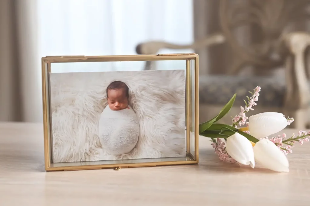 A framed photo of a swaddled newborn baby lying on a soft, white furry blanket sits on a wooden surface next to three white tulips and pink flowers, with blurred furniture in the background. Kristin Milito Photography - Newborn & Maternity Photographer - Chicago IL