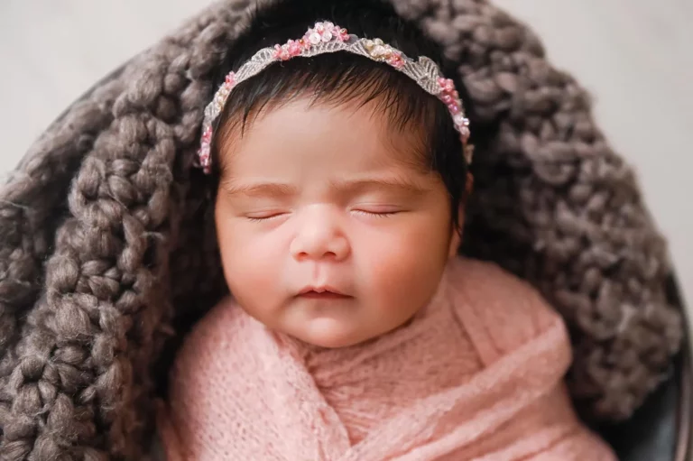 A close-up of a sleeping baby wrapped in a soft pink blanket, resting on a large gray knitted blanket. The baby has dark hair and is wearing a delicate headband with pink and clear gemstones. - Image by Kristin Milito Photography, Chicago IL.