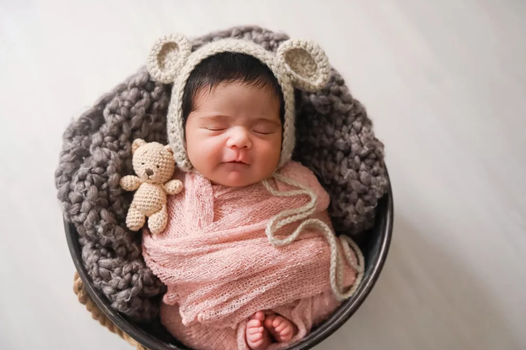 A newborn baby, swaddled in a pink blanket, sleeps peacefully in a round basket lined with a gray chunky knit blanket