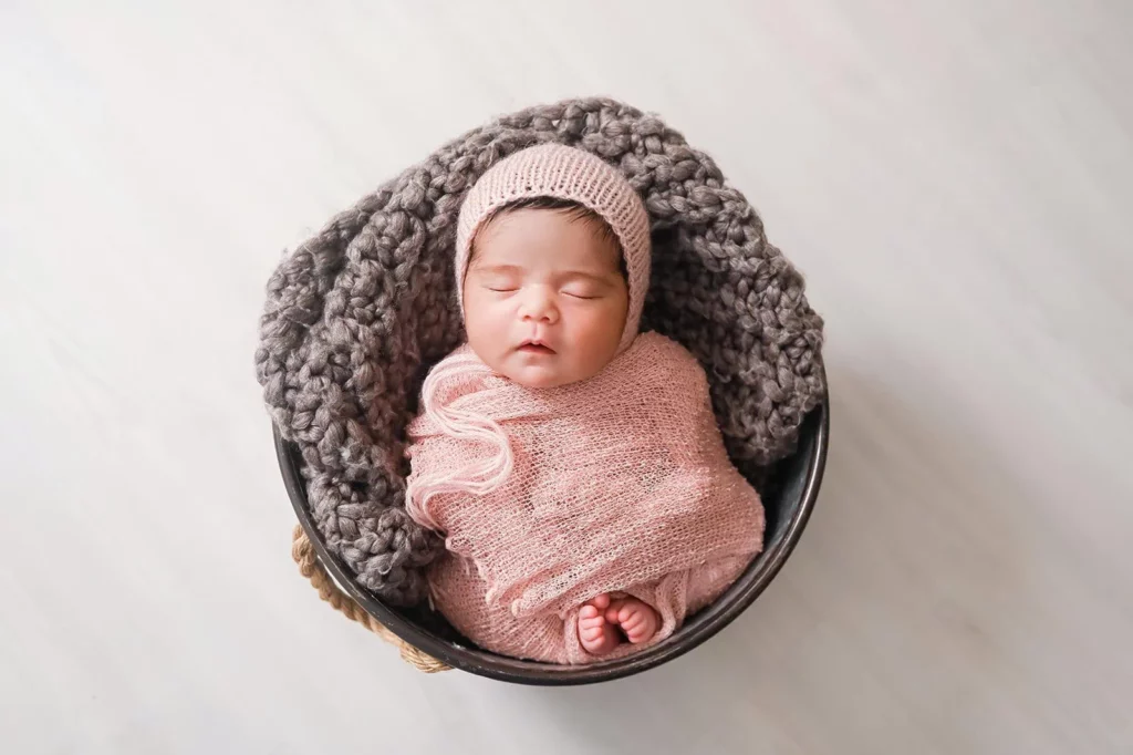 A newborn baby peacefully sleeps, swaddled in a soft pink blanket, with a matching pink knit hat. The baby rests in a round, dark basket lined with a chunky grey knit blanket. The background is a soft, neutral tone. - Image by Kristin Milito Photography, Chicago IL.