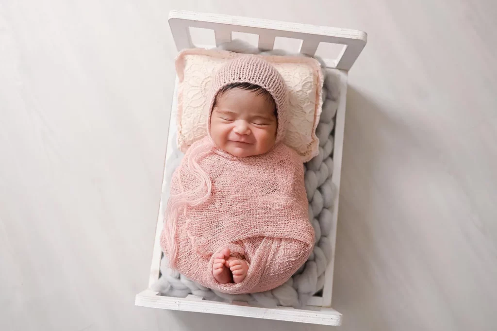A newborn baby, swaddled in a pink knit wrap and wearing a matching hat, is sleeping peacefully in a white wooden crib. The baby's face shows a gentle smile, and the crib is lined with a soft, textured blanket. The background is light grey. - Image by Kristin Milito Photography, Chicago IL.