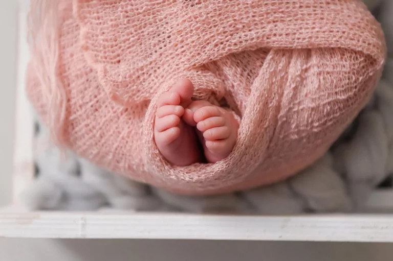 Close-up of a newborn baby's feet peeking out from a pink, textured blanket. The baby is wrapped snugly in the blanket, and the background is soft and neutral, creating a cozy and serene atmosphere. - Image by Kristin Milito Photography, Chicago IL.