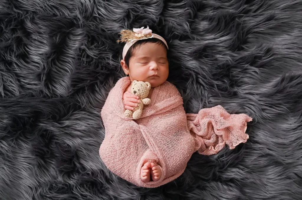 A newborn baby sleeps peacefully on a dark, fluffy surface. The baby is wrapped in a soft pink cloth, holding a small teddy bear, and wears a delicate headband with a tiny decorative flower. The overall scene is calm and serene. - Image by Kristin Milito Photography, Chicago IL.