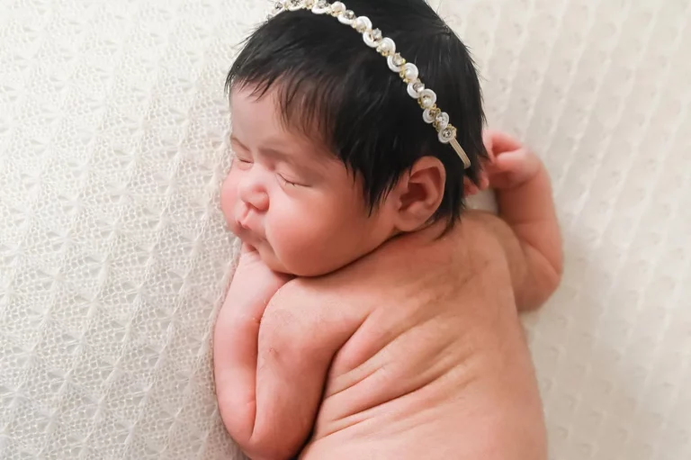 A sleeping newborn baby with dark hair wears a delicate pearl headband. The baby is posed on their side against a soft, textured white background, accentuating the peaceful and serene nature of the scene.