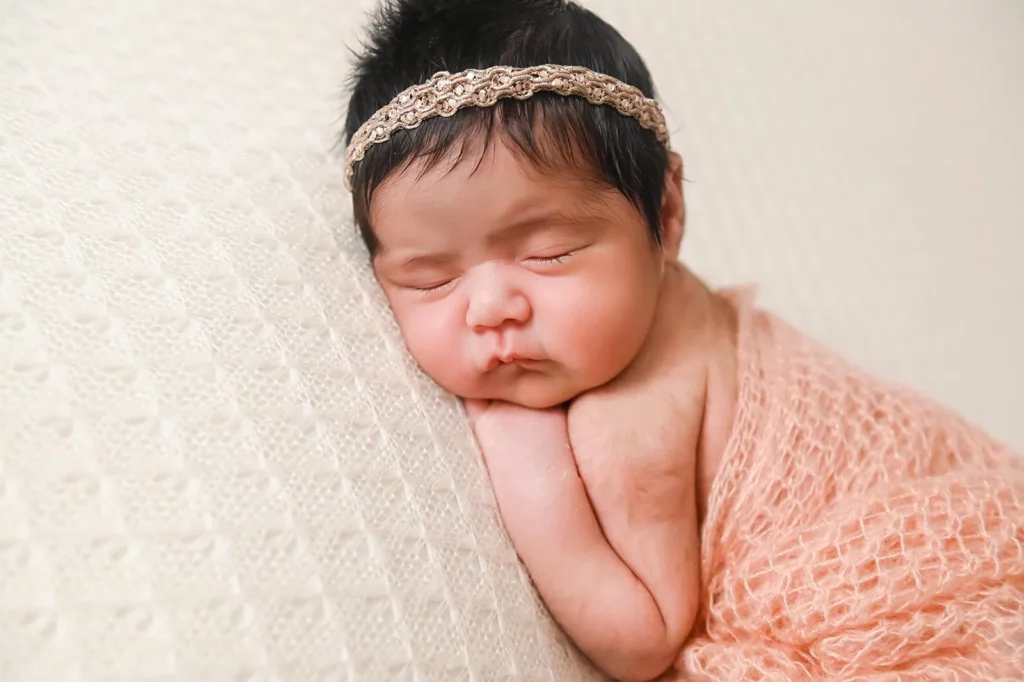A newborn baby with dark hair is peacefully sleeping while propped up on a soft, textured blanket