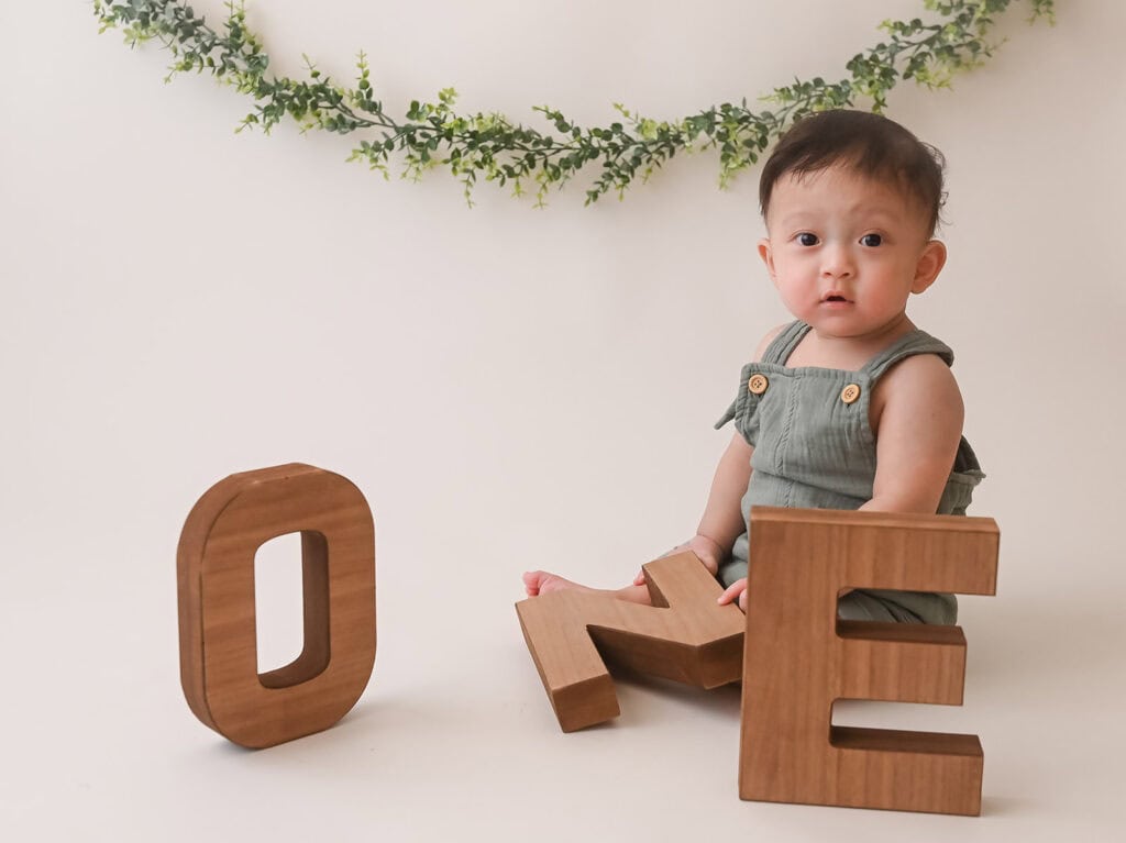 Baby in green outfit sitting with wooden letters 'ONE'.