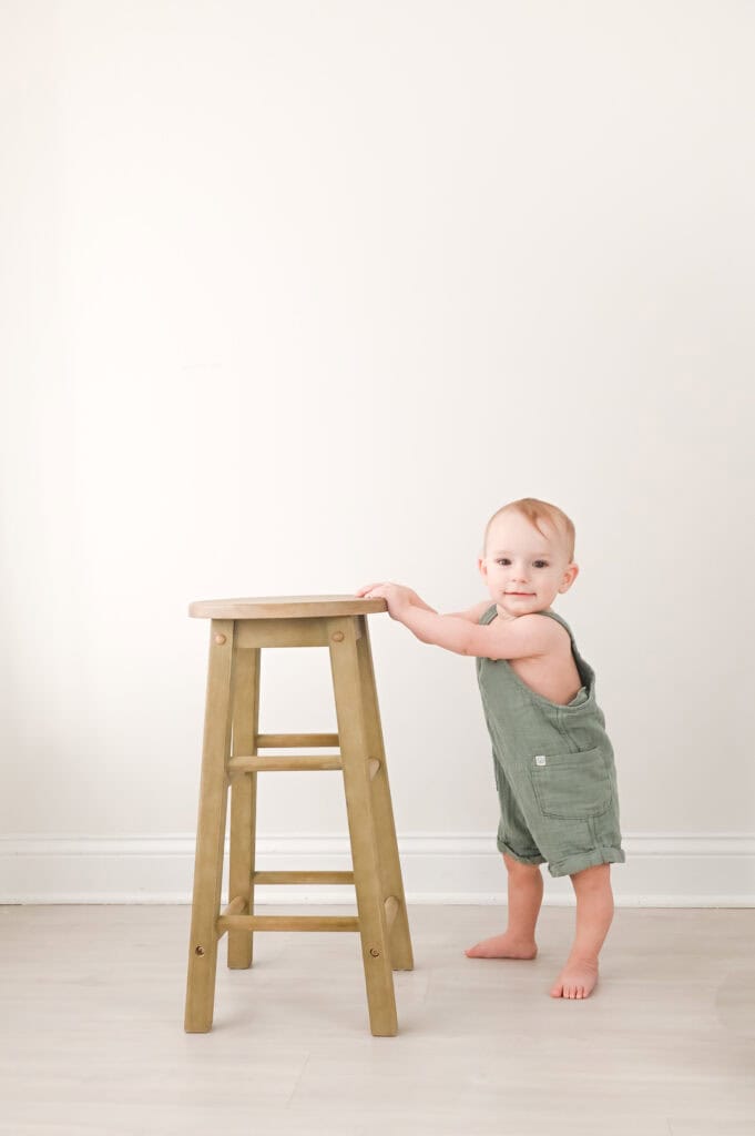 Baby standing beside wooden stool indoors.